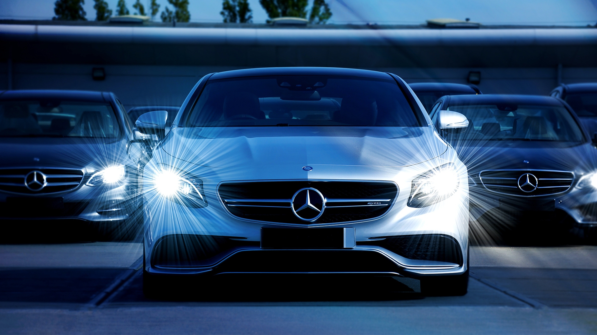 Front view of a silver Mercedes-Benz with headlights on, surrounded by other luxury cars at night.