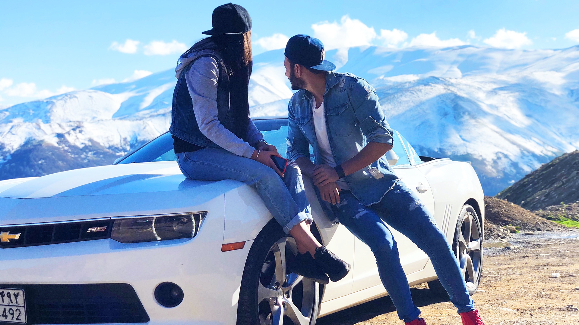 Couple sitting on the hood of a white Chevrolet Camaro, enjoying a scenic snowy mountain view.