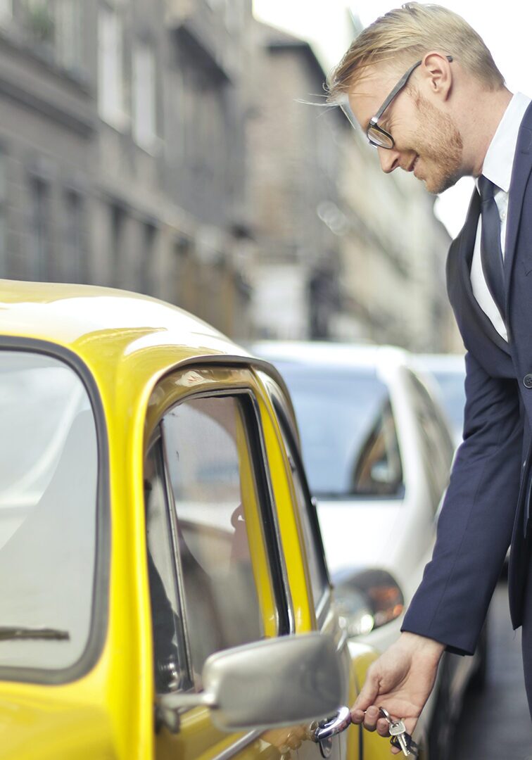 Man in a business suit unlocking a yellow vintage car on a city street.